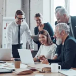 group sitting and standing around a table looking at a laptop collaborating